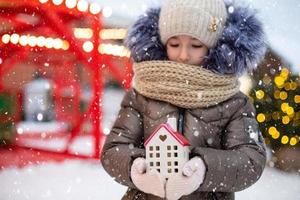 casa, casa de campo em miniatura nas mãos da garota usando luvas e roupas quentes ao ar livre na neve. valores familiares, compra de habitação, realocação, hipoteca. casa aconchegante, natal, reserva de ano novo foto