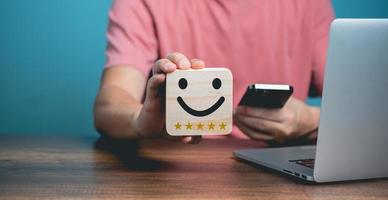 conceito de satisfação de atendimento ao cliente, jovem empresário usando telefone celular e segurando o rosto feliz de madeira com rosto sorridente, ícone de classificação de cinco estrelas de satisfação. foto