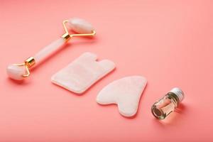 um conjunto de ferramentas para a técnica de massagem facial gua sha feita de quartzo rosa natural em um fundo rosa. rolo, pedra de jade e óleo em uma jarra de vidro para cuidados com o rosto e corpo.
