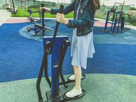 fazendo esportes no simulador no parque ao ar livre. uma garota de vestido azul e tênis branco treina as pernas e os braços na plataforma. lazer ativo, tempo ao ar livre foto