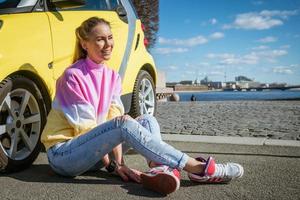 uma jovem feliz está sentada no asfalto encostada em um carro amarelo em um dia ensolarado de primavera foto