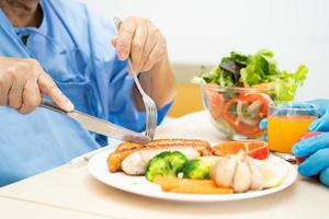 paciente idosa asiática ou idosa tomando café da manhã e comida saudável vegetal com esperança e feliz enquanto está sentado e com fome na cama no hospital. foto