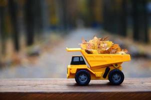 um pequeno caminhão amarelo de brinquedo é carregado com folhas amarelas caídas. o carro fica em uma superfície de madeira contra um fundo de um parque de outono embaçado. limpeza e remoção de folhas caídas. trabalhos sazonais foto