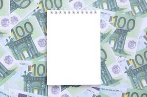 caderno branco com páginas limpas sobre um conjunto de denominações monetárias verdes de 100 euros. muito dinheiro forma uma pilha infinita foto