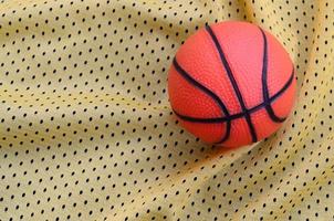 pequena bola de basquete de borracha laranja encontra-se em uma textura de tecido de roupa de camisa esportiva amarela e fundo com muitas dobras foto