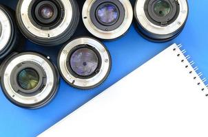várias lentes fotográficas e caderno branco estão sobre um fundo azul brilhante. espaço para texto foto