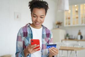 feliz menina adolescente de raça mista segurando o cartão de crédito do banco, compras no smartphone. serviços bancários on-line foto