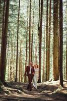 feliz casal elegante recém-casados na floresta verde em dia de verão. noiva de vestido longo branco e noivo de terno vermelho estão abraçando. dia do casamento. foto