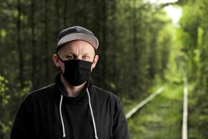 jovem com máscara antiviral protetora preta no parque de verão. o cara está descansando ao ar livre em um dia ensolarado de verão em uma máscara feita por suas próprias mãos. Proteção contra vírus. medidas de quarentena foto