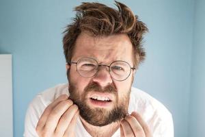 homem desgrenhado toca sua barba, ele está perplexo, insatisfeito e irritado com sua aparência desarrumada, de pé contra uma parede azul. foto