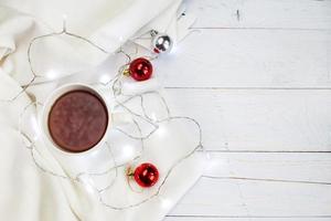 xícara de chá ou café, manta branca de malha, guirlanda e bolas de natal em fundo de madeira. conceito de manhã de natal foto