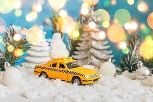 fundo de bandeira de natal. modelo de táxi de carro de brinquedo amarelo e ornamentos de decorações de inverno em fundo azul com neve e luzes de guirlanda desfocadas. conceito de serviço de táxi de entrega de tráfego da cidade.