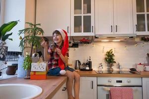uma criança com um chapéu de papai noel está sentada em uma cozinha festiva decorada para o natal. uma garota feliz veste uma araucária de planta de casa como uma árvore de natal. ano Novo foto