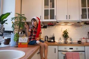 uma criança com um chapéu de papai noel está sentada em uma cozinha festiva decorada para o natal. uma garota feliz veste uma araucária de planta de casa como uma árvore de natal. ano Novo foto