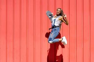 mulher negra vestida casual, com um skate pulando de felicidade no fundo da parede urbana vermelha. foto