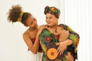 mãe e filha mais velha em vestido tradicional estilo nativo africano carregam bebê recém-nascido de um mês de idade. afro-asia de raça mista infantil feliz durma bem no abraço caloroso da maternidade. foto