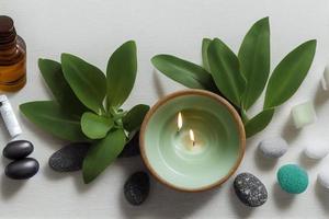 itens de tratamento de beleza para procedimentos de spa na mesa de madeira branca com planta verde. pedras de massagem, óleos essenciais e sal marinho com vela acesa. foto