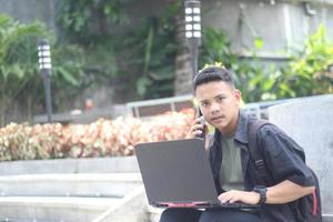 atraente jovem asiático usando laptop no espaço de coworking com cara feliz e chamando sua amiga foto