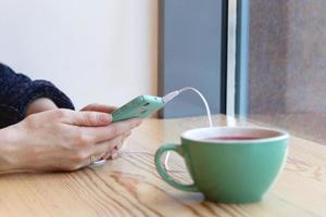 mãos de jovem com celular azul e xícara de chá em uma mesa em um café. foto