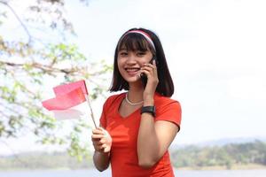 bela jovem asiática carregando a bandeira indonésia com um rosto alegre e chamando seu amigo foto