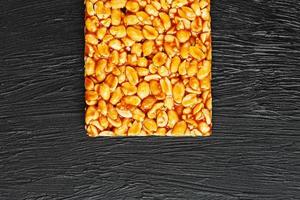 uma grande telha dourada de amendoim, uma barra em um melaço doce em um fundo de textura preta. kozinaki doces úteis e saborosos do leste foto