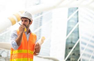 homem caucasiano rengineer usa um walkie-talkie para falar, vestindo colete laranja e grande capacete, e a outra mão segurando a planta branca no local de trabalho do centro da cidade. foto