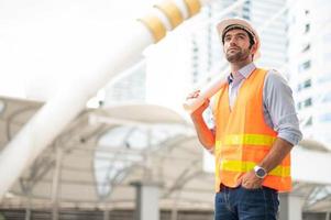 jovem homem caucasiano segurando um papel grande, cara vestindo camisa azul clara e jeans com colete laranja e capacete branco para segurança na área de construção. foto