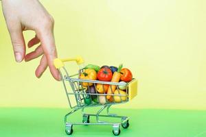 mão segurando o carrinho de compras com frutas e legumes em fundo verde e amarelo. conceito de entrega de comida foto