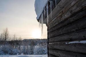 pingentes de gelo pendem do telhado coberto de neve de um banho de madeira, tendo como pano de fundo uma paisagem de inverno, no campo. foto