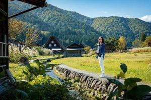 linda garota japonesa com um fundo de aldeia de shirakawago durante o outono com uma casa de triângulo, campo de arroz e montanha de pinheiros. foto