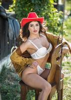 mulher morena sensual com look country sexy na cadeira. retrato de uma menina com lingerie branca e chapéu vermelho. garota se divertindo com botas de couro marrom no rancho. estilo country sexy americano foto