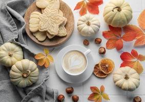 café quente com biscoitos em um copo branco cercado por folhas de outono e abóboras em um fundo branco de madeira.