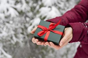 presente na caixa verde com fita vermelha nas mãos femininas contra abeto nevado no fundo de natal da floresta de inverno e presentes de feriados de ano novo foto