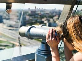 uma linda garota olha para a paisagem, um panorama da cidade na sala de observação, um par de binóculos, um telescópio com um aceitador de notas em um deck de observação ao ar livre foto