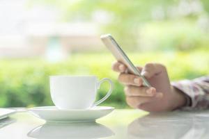 mãos masculinas segurando um telefone inteligente e uma xícara de café na mesa foto