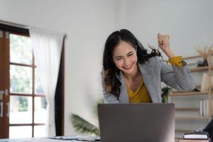 feliz mulher asiática animada comemorar com computador portátil, sucesso ou ação feliz. freelancer ou empreendedor usando tecnologia e iniciando uma casa moderna. foto