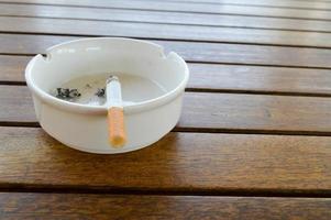 um cinzeiro de cerâmica branca com um cigarro e cinzas com um isqueiro preto para fumar fica sobre uma mesa de madeira em um café foto
