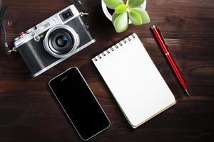 câmera clássica com página de bloco de notas em branco e caneta vermelha na mesa de madeira marrom escura, mesa vintage com telefone e flor verde foto