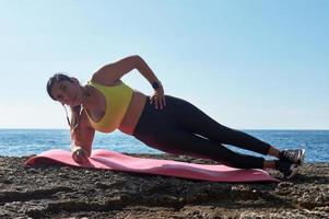 mulher latina, meia-idade, vestindo roupas esportivas, treinando, fazendo exercícios físicos, prancha, abdominais, degrau de alpinista, queimando calorias, mantendo a forma, ao ar livre à beira-mar, usando fones de ouvido, relógio inteligente foto