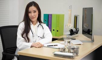 retrato de sorridente médica asiática vestindo jaleco branco com estetoscópio sentado atrás da mesa no escritório. foto