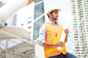 jovem caucasiano segurando um papel grande, cara vestindo camisa azul clara e jeans com colete laranja e capacete branco para segurança na área de construção. foto