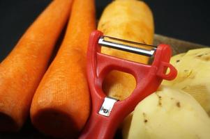 descascador vermelho com cenouras descascadas e batatas isoladas em fundo escuro e preto foto