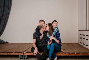 família feliz com dois filhos, em roupas casuais, é fotografada sentada no chão. photoshoot de uma família com 2 filhos, fundo branco. relações familiares calorosas. beijo mãe e pai foto