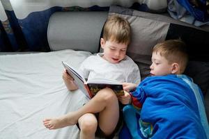 irmãozinho lê um livro na cama para um ancião. amizade entre irmãos. amor pela leitura. cotovia e coruja entre as pessoas. 2 meninos na cama estão lendo um livro. histórias de ninar foto