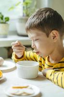 menino toma café da manhã com cereais com leite. café da manhã na cozinha antes da escola. o menino está comendo na mesa perto da janela. foto