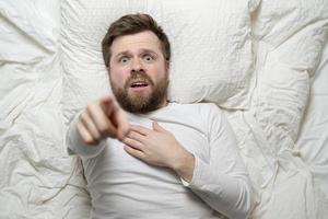 homem assustado deita na cama apontando para cima e segura a mão no peito, ele está agitado, teve um pesadelo. conceito de saúde psicológica. foto