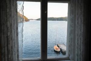 velha janela fechada com excelentes vistas sobre o mar, costa rochosa, barco atracado e céu ao pôr do sol. foto