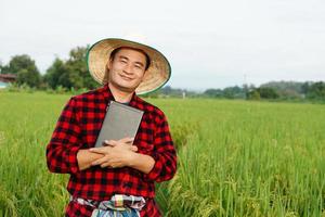 homem agricultor asiático está no arrozal, usa chapéu e camisa xadrez vermelha, segura tablet inteligente para inspecionar o crescimento e as doenças das plantas. conceito, agricultor inteligente, use tecnologia na agricultura. foto