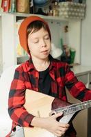 menino de chapéu vermelho e camisa xadrez toca balalaica. menino bonito segurando seu violão. aulas de música em casa. passatempo para a alma. ensino de musica em casa foto