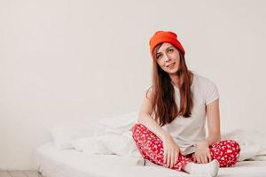 garota engraçada sorridente em um chapéu vermelho e pijama de natal, sentado na cama com lençóis brancos. feriados para o natal. garota de bom humor natalino. garota de camiseta branca e pijama, contra foto